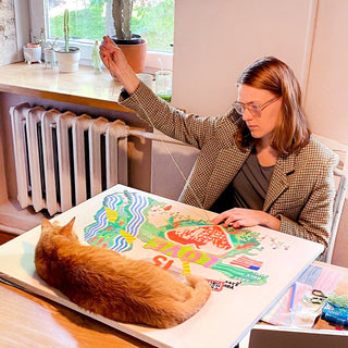 Lithuanian illustrator Daukantė drawing at her desk while her orange cat Bosch sleeps on the paper
