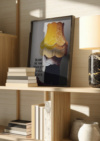 Solsken by Grafik bageri in a dark frame on a wooden shelf beside books and a lamp