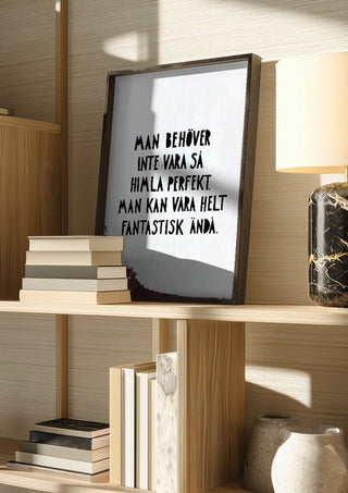 Fantastisk by Grafik bageri in a dark frame on a wooden shelf beside books and a lamp