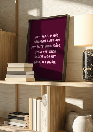 Modig by Grafik bageri in a dark frame on a wooden shelf beside books and a lamp