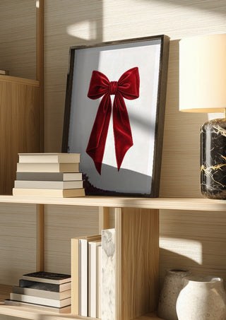 Red velvet bow by Ohkimiko in a dark frame on a wooden shelf beside books and a lamp