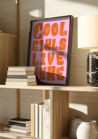 Cool girls live here by Ohkimiko in a dark frame on a wooden shelf beside books and a lamp