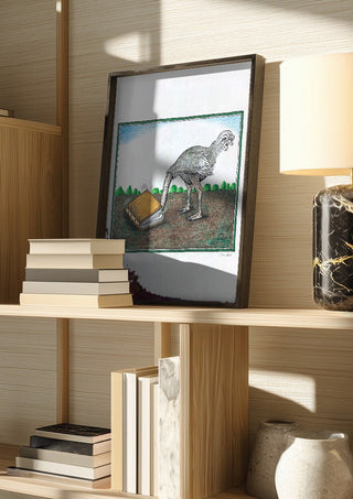 The Ostrich and the Book by Waldemar Mandzel in a dark frame on a wooden shelf beside books and a lamp