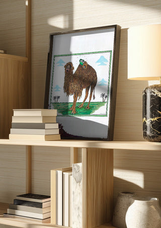 The Camel and the Book by Waldemar Mandzel in a dark frame on a wooden shelf beside books and a lamp