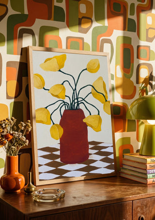 Gula blomster i terracottafärgad vas by Rebecka Hansson in a wooden frame on a sideboard against a retro patterned wall