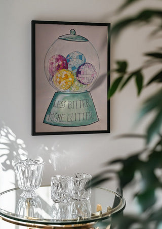 Less Bitter More Glitter by L.OE. Art in a black frame above a glass table with crystal glassware