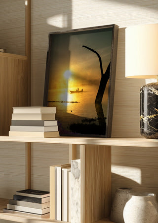 Fishermen Of Naivasha by Gloria Mwivanda in a dark frame on a wooden shelf beside books and a lamp