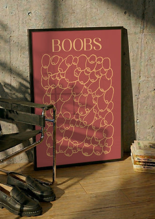 Boobs by Astrid Lind in a black frame leaning against a concrete wall by a steel chair