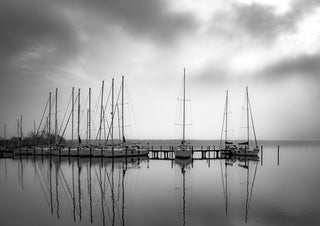 Warten auf den Wind by Guido Lucker - black and white photograph of sailing boats at a jetty reflected in calm water