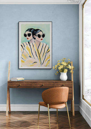 Framed "Yellow Twins" by David Wolf above a wooden desk with chair and vase of yellow flowers in a modern room.